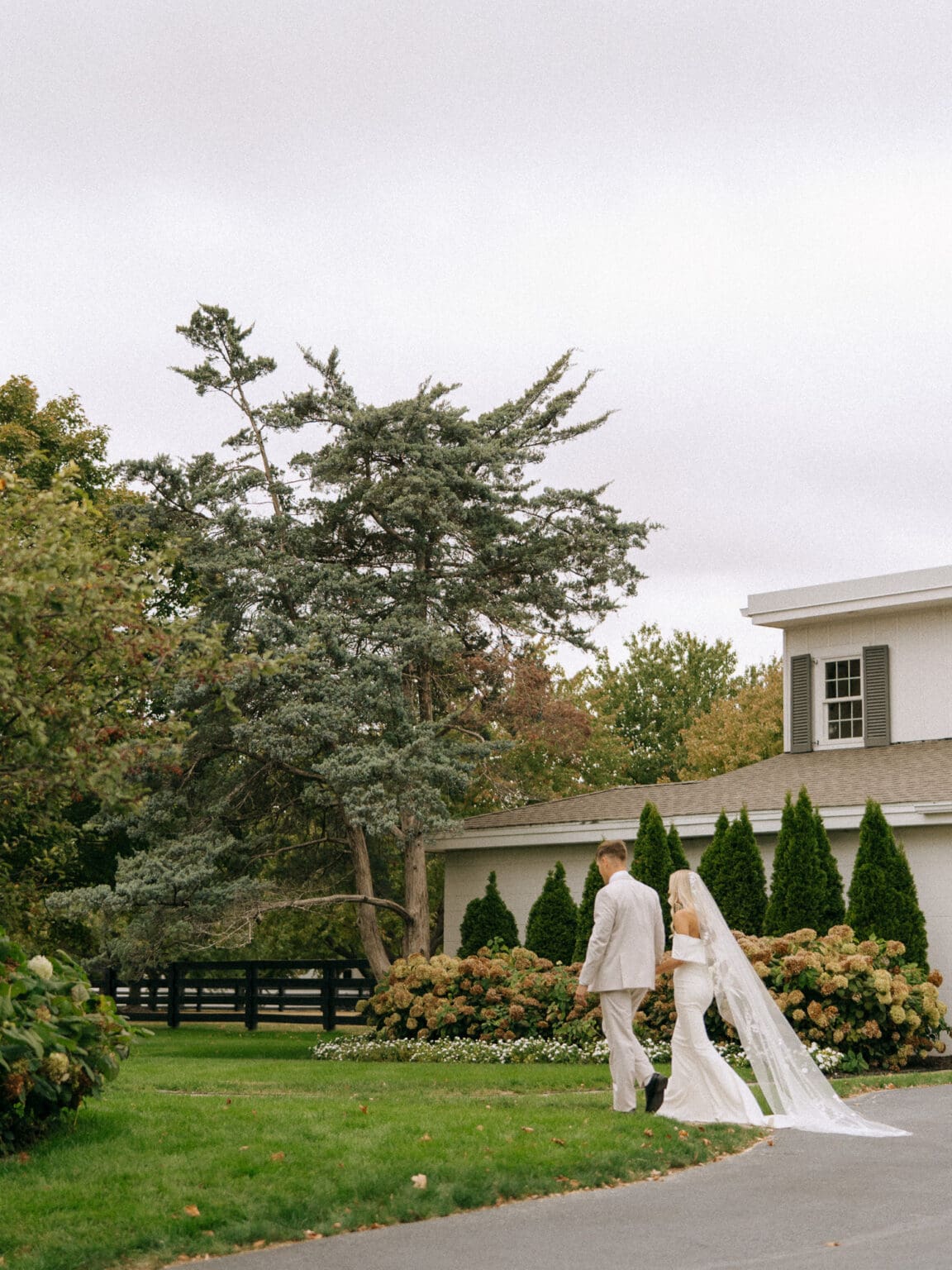 groom and bride walking through grass and trees near white building