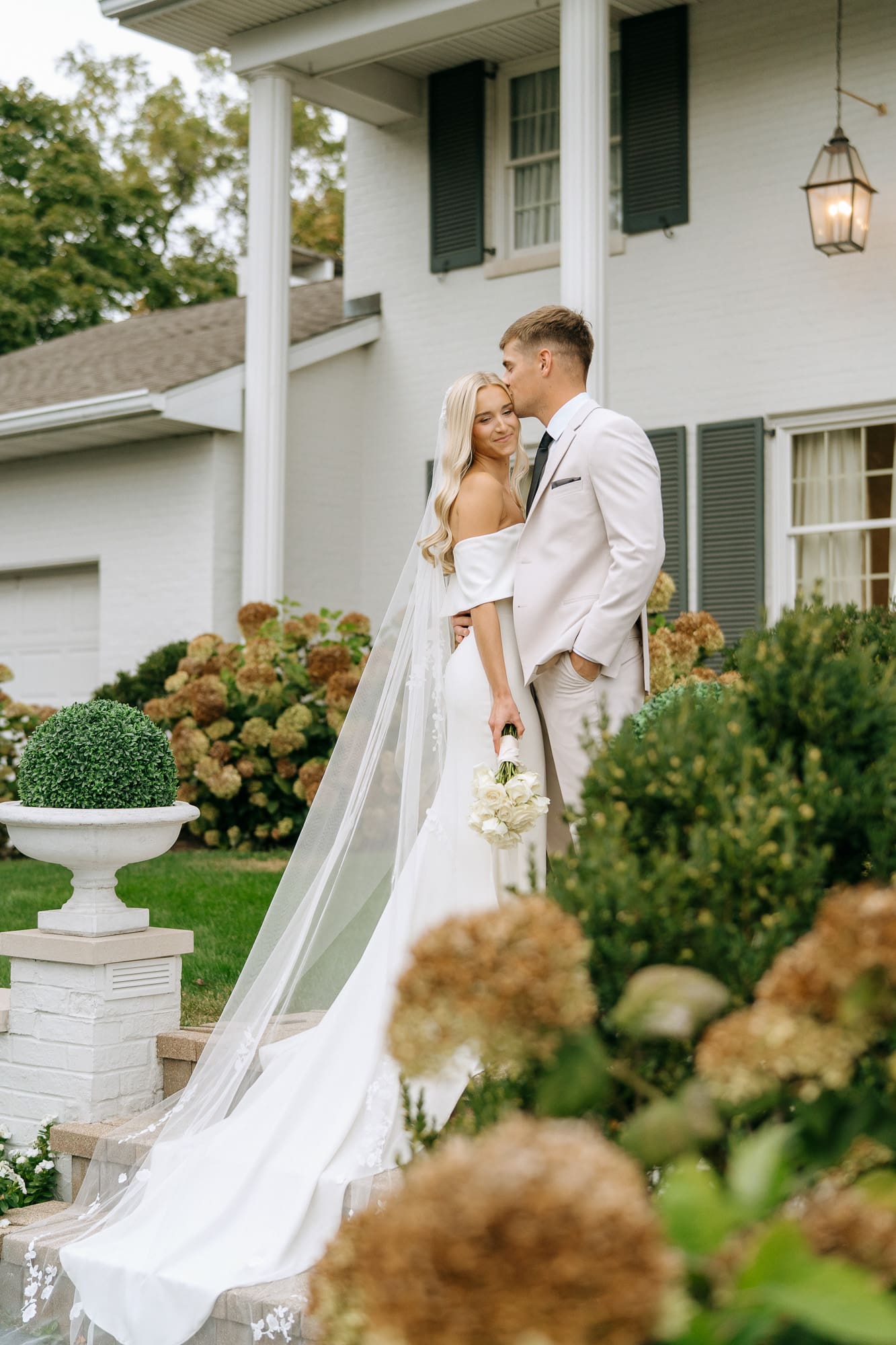 bride and groom standing on stairs in front of white house, with green bushes