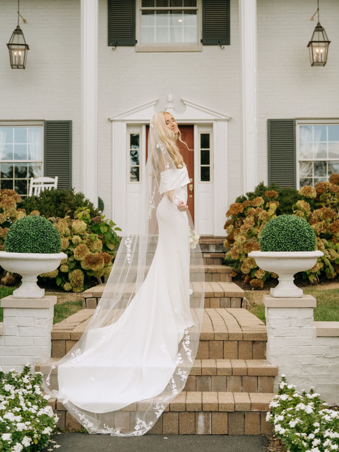 Bride wearing long white veil and standing on stairs in front of white house